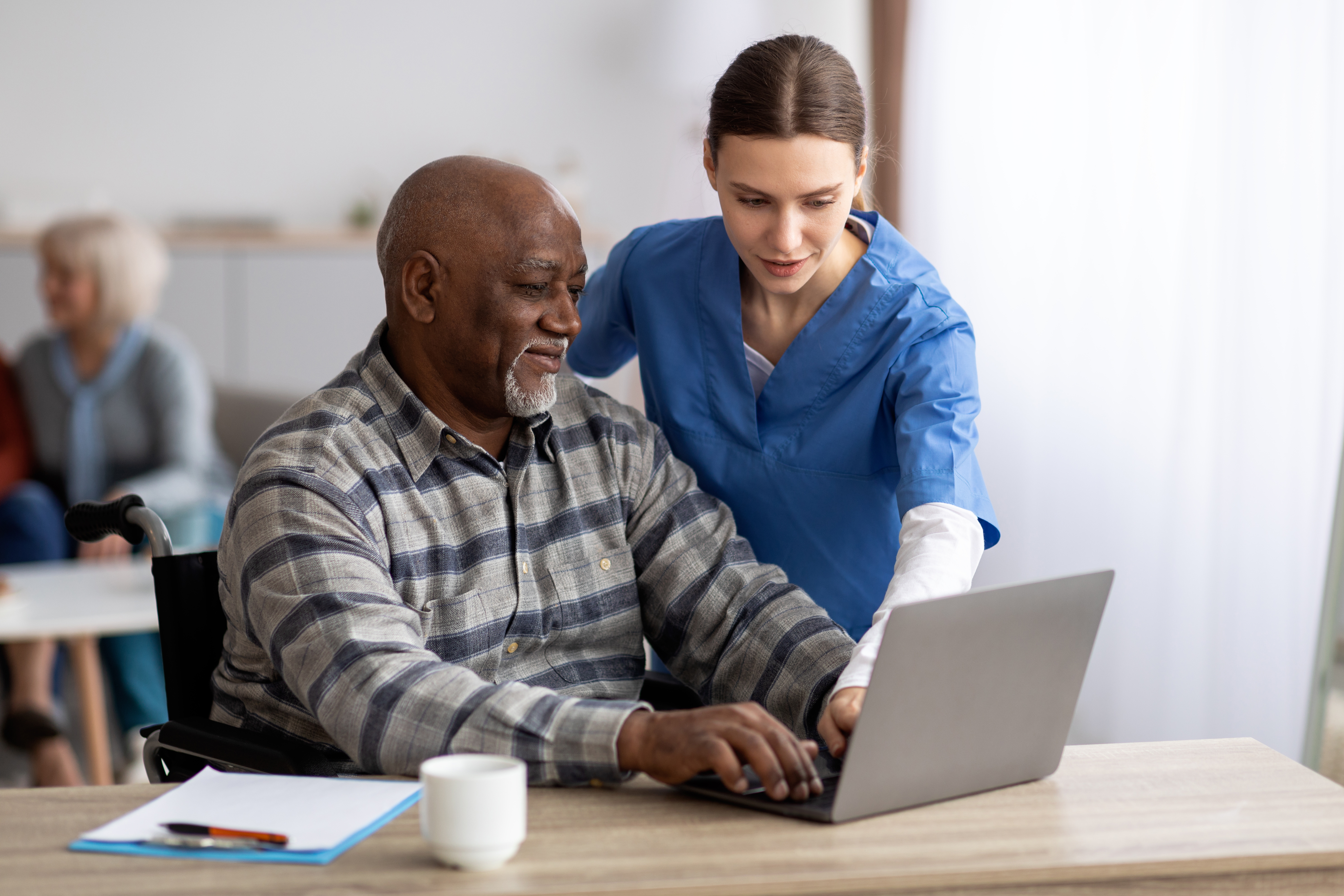 Friendly young woman nurse helping black senior man in wheelchair using laptop, smiling elderly African American male patient surfing on Internet at nursing home, copy space