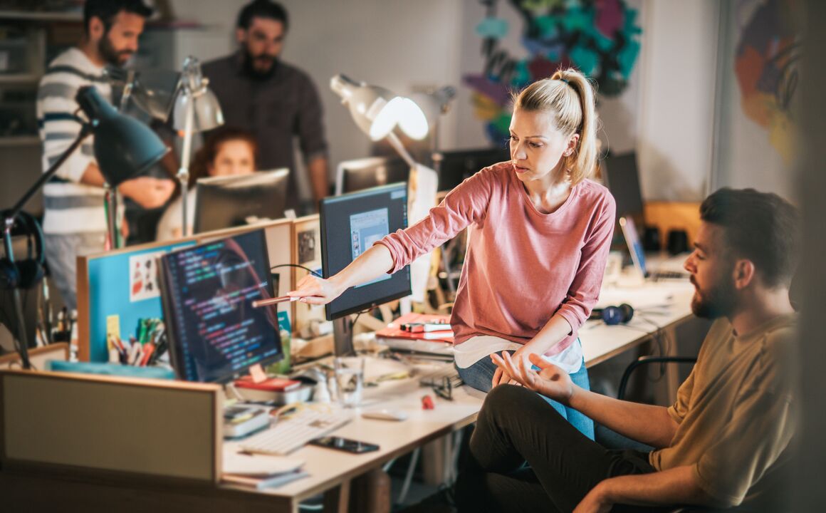 Woman sitting on her collegaues desk point at his computer monitor