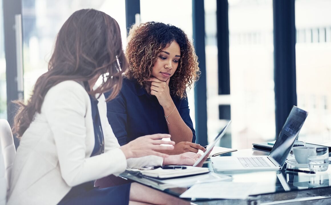 Two professional women discuss UI program efficiencies while looking at a tablet