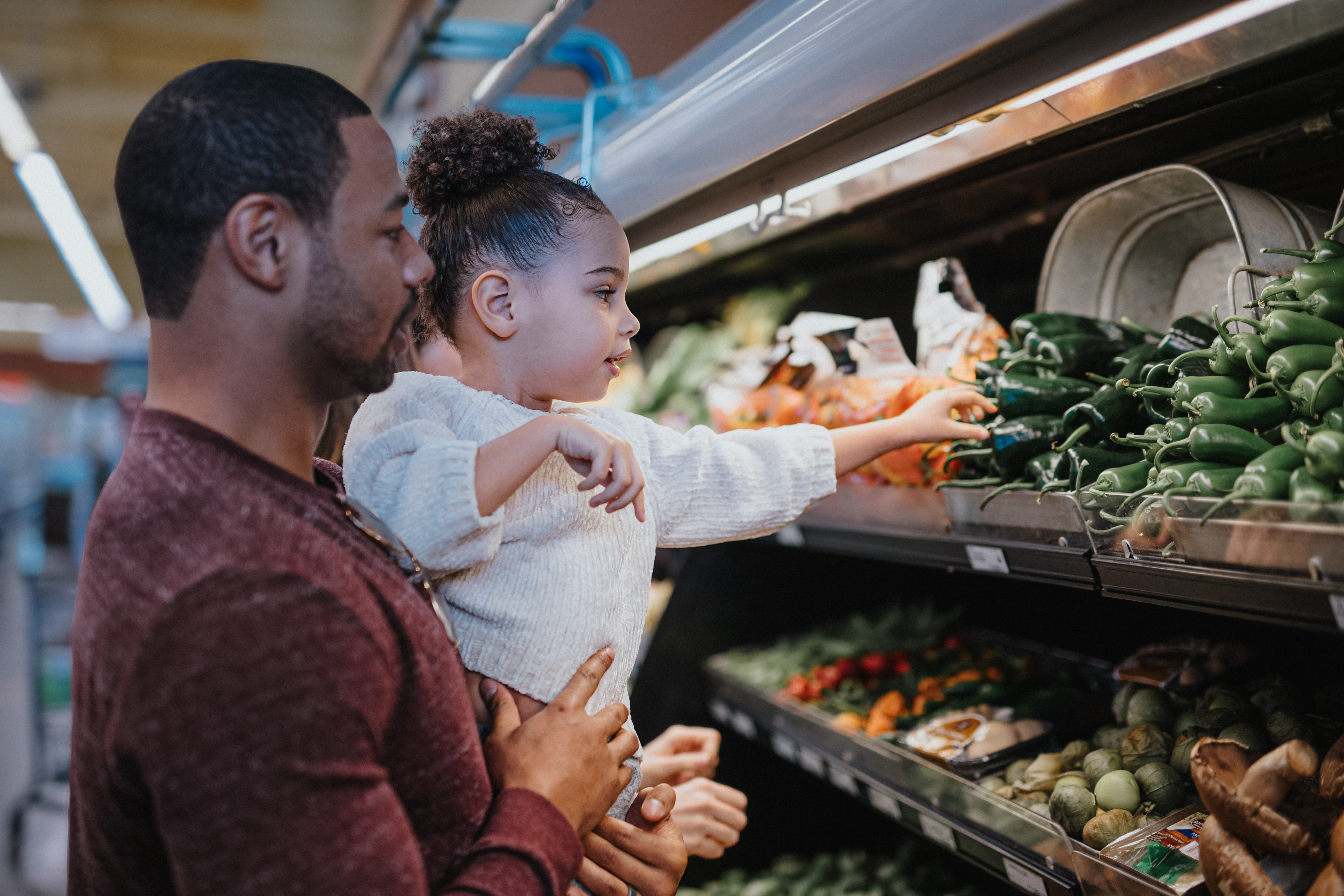 A loving father of African American descent holds his young daughter so she can pick out vegetables at the grocery store.