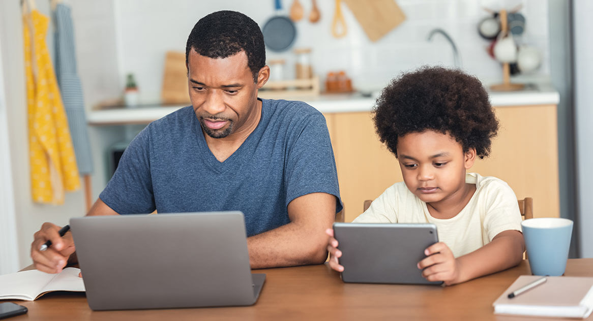 Father works on laptop while toddler son views a tablet while sitting at the kitchen table.
