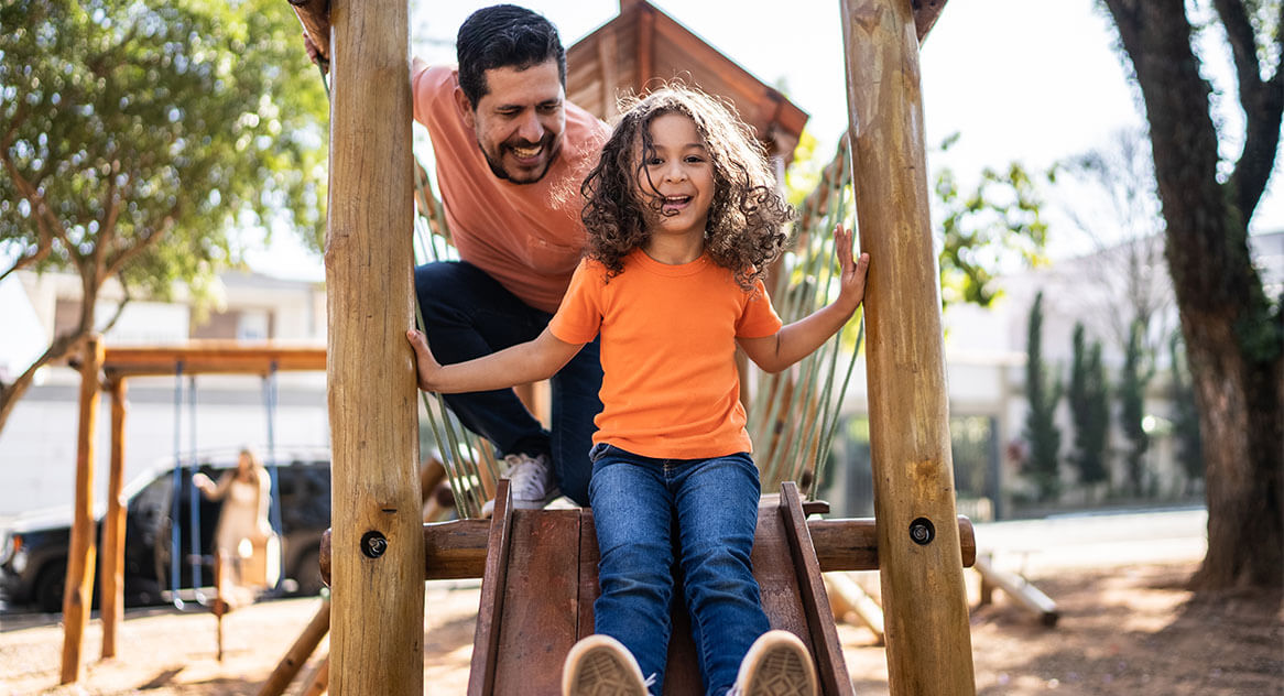 Father pushes daughter down the slide of a wooden playground structure.