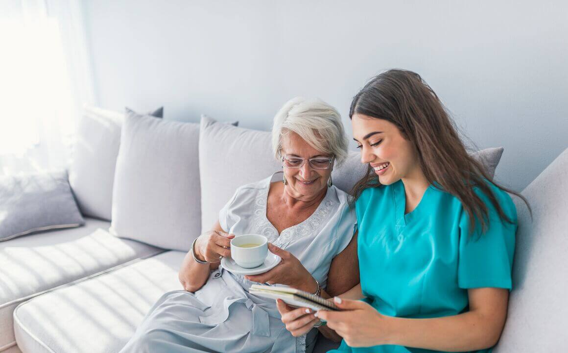 Happy patient is holding caregiver for a hand while spending time together. Elderly woman in nursing home and nurse. Aged elegant woman and tea time at nursing home