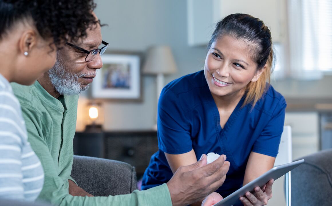 Home health worker holding a tablet smiles at older couple seated on couch