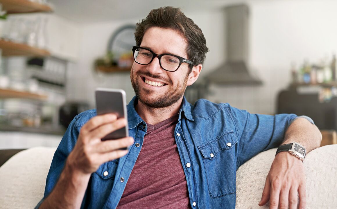 Smiling young man looks at phone while sitting on couch at home