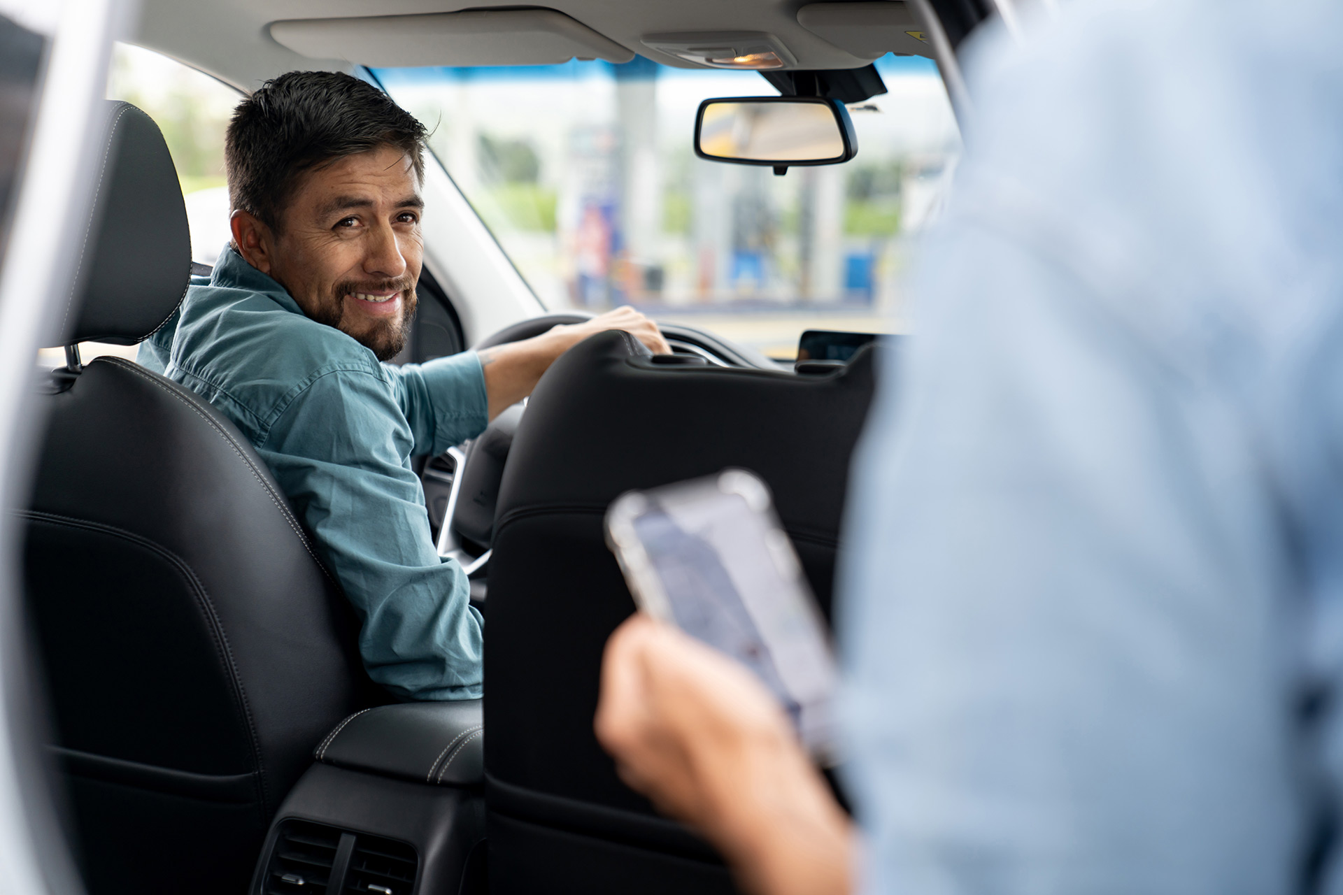 A noncustodial father, who earns income by driving for an on-demand rideshare service, turns and smiles at a passenger entering the back seat of his vehicle. The father is wearing a long-sleeve green shirt, the interior of the vehicle is black leather, and the passenger is holding a mobile phone. 