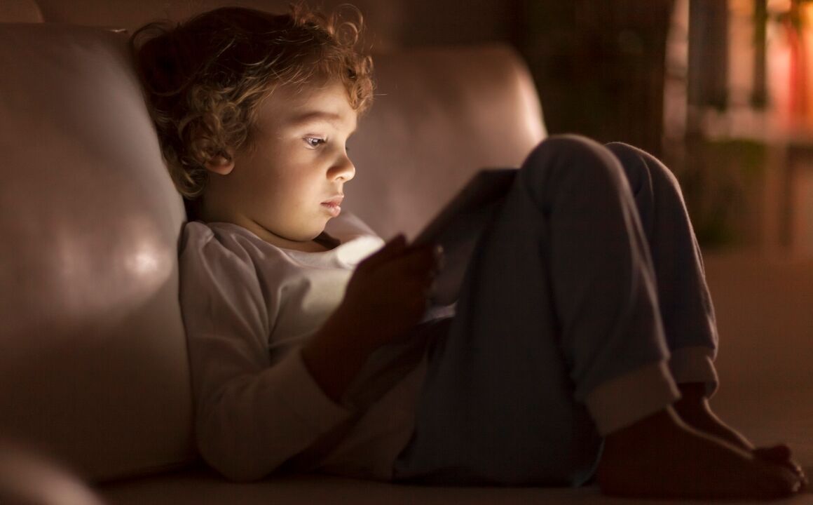 A young boy sits on a couch with his legs drawn up to him and his expression neutral. The room is dark, but the tablet on his lap illuminates his face.