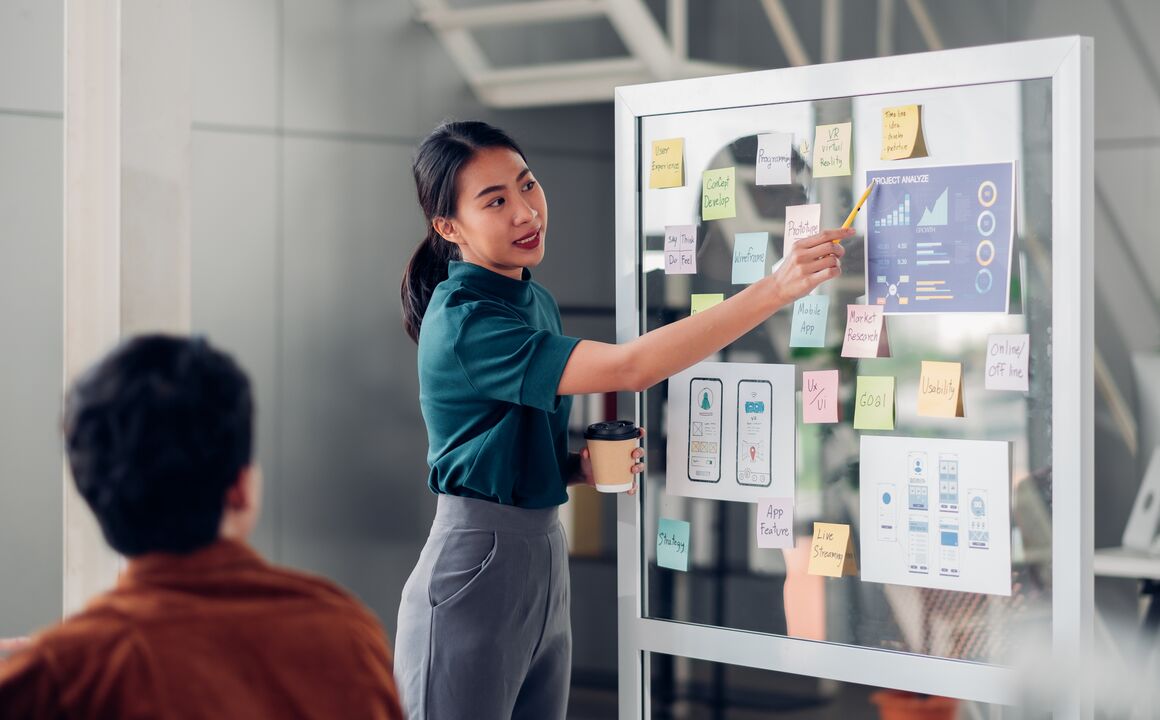 Female web accessibility expert points to timelines and strategies on a whiteboard while presenting a digital accessibility roadmap to a male chief information officer sitting in front of her.  