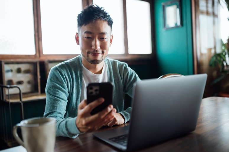 Young man enrolls in health coverage using the smartphone in his hands while sitting at a table with a laptop and coffee mug.