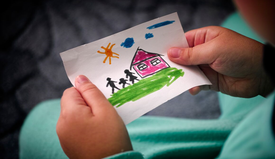 Child's hands hold a marker drawing of a house, three people, and a sun