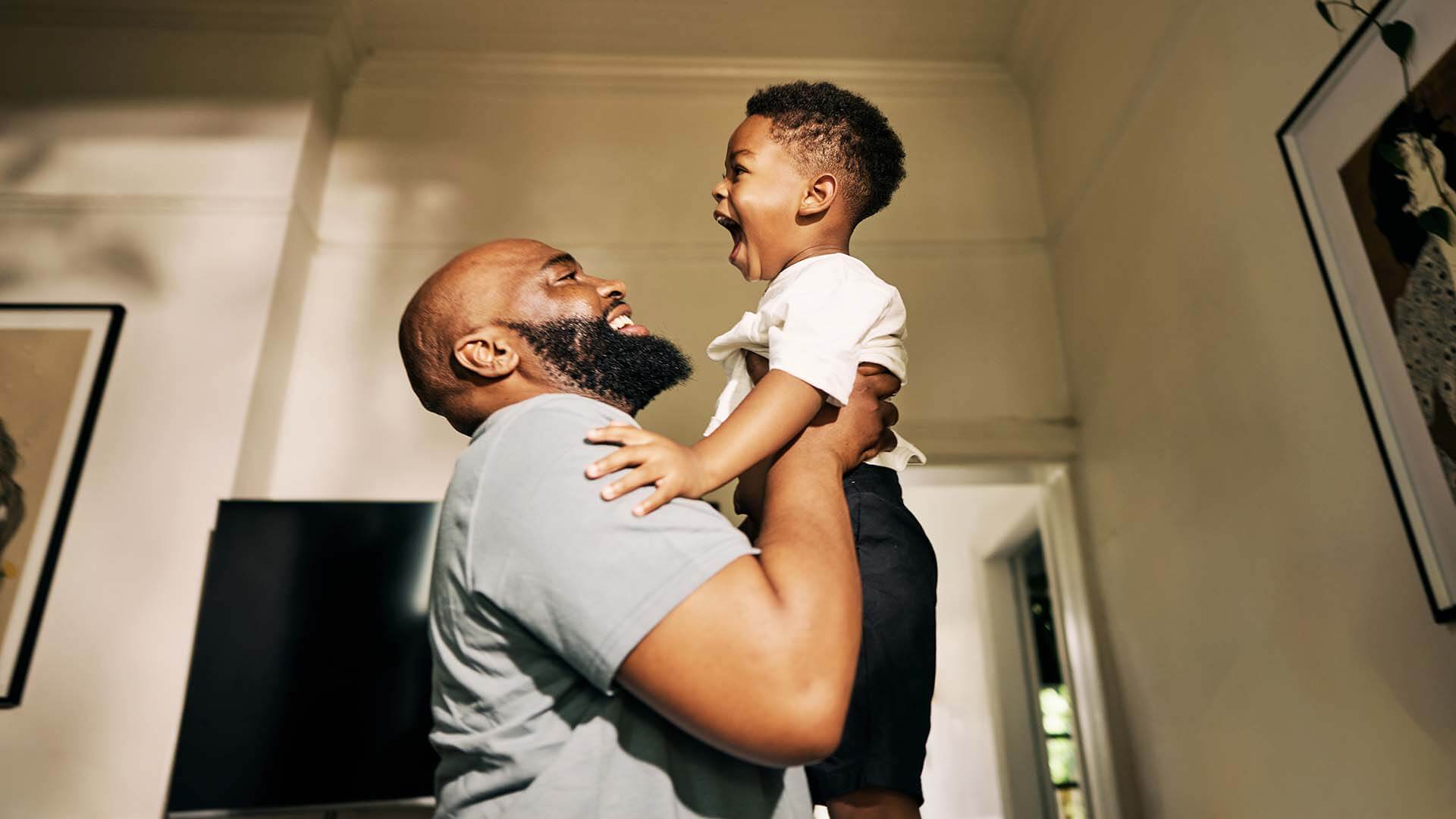 A non-custodial father celebrates his new job by lifting his toddler into the air of a sun-dappled room.