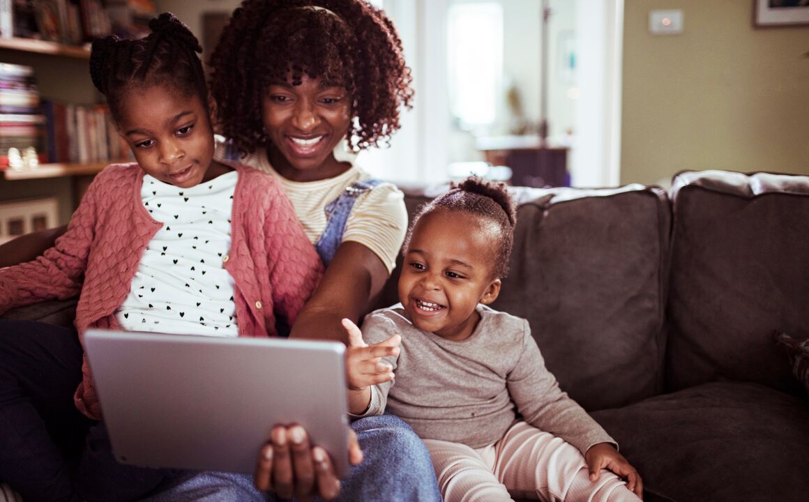 A mother sits on the couch and snuggles with her two young daughters while using a mobile app on her tablet to upload health insurance documents. They are all smiling and looking at the screen.    