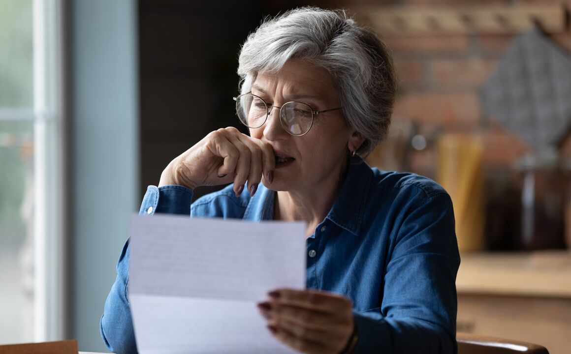 A woman sits at her kitchen table as she reviews a letter about meeting new Medicaid work requirements and continuing her coverage. One hand is holding the letter in front of her, the other hand is touching her mouth as she concentrates. 
