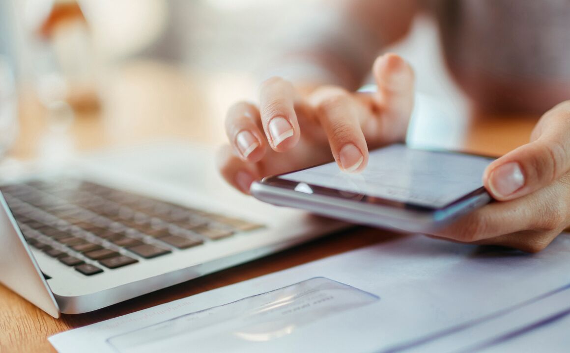 A close-up of a woman’s hands as she holds a mobile phone over a stack of papers. She is tapping on the screen to scan and upload a document for health insurance enrollment.
