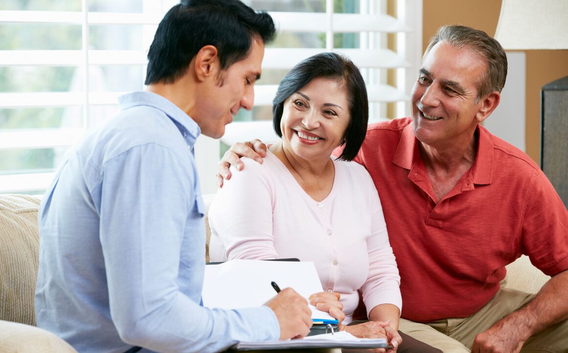 A smiling senior woman participates in an independent assessment in her home. She is seated on a couch, between the assessor taking notes and her husband with his arm around her shoulders. 