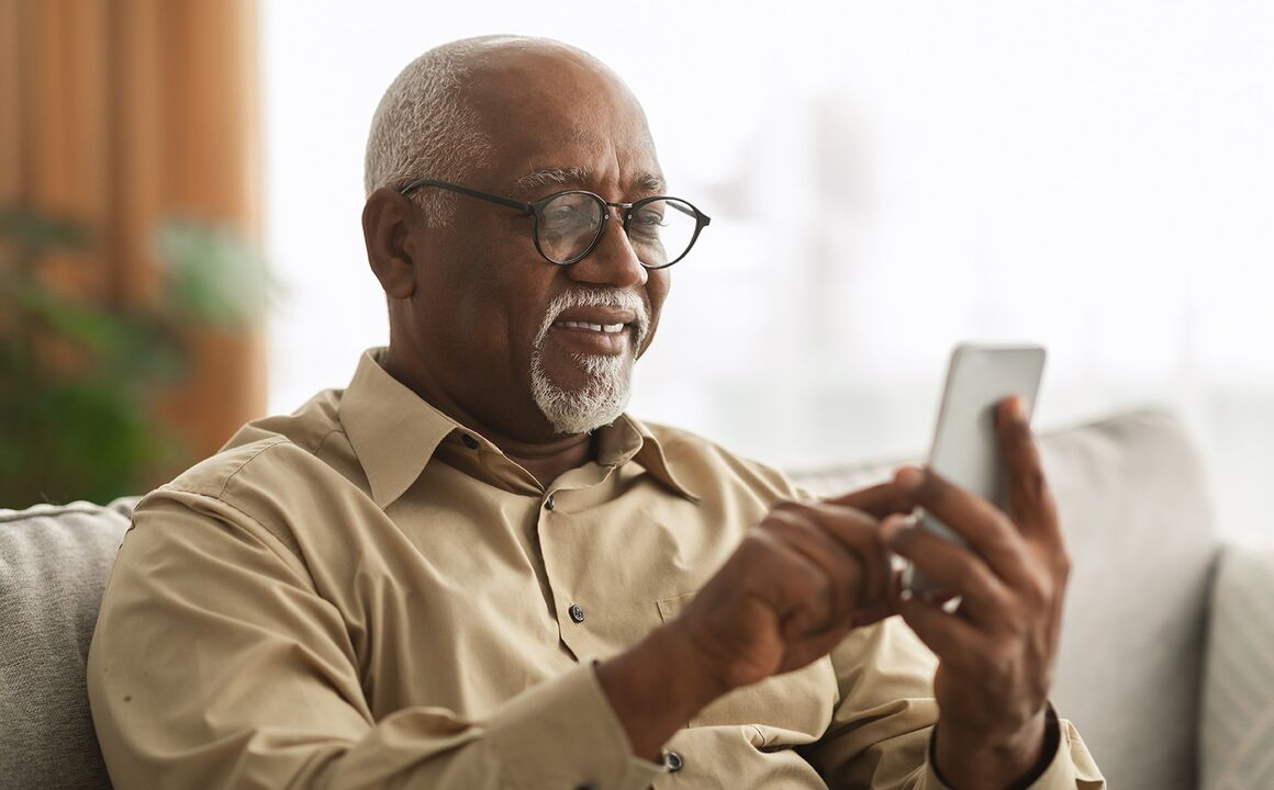 Senior man uses his smartphone to contact the California LifeLine program to inquire about lowering his cell phone bill. He is wearing eyeglasses and sitting on a couch at home.