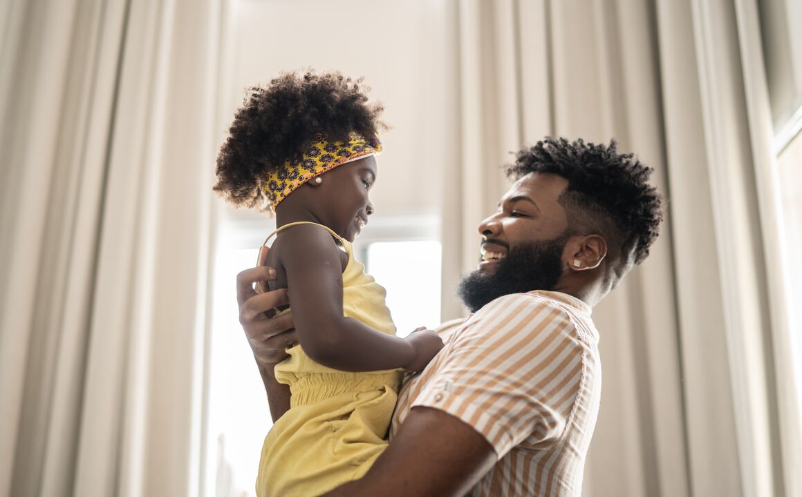 A father embraces his toddler daughter in front of a window in their new home. 