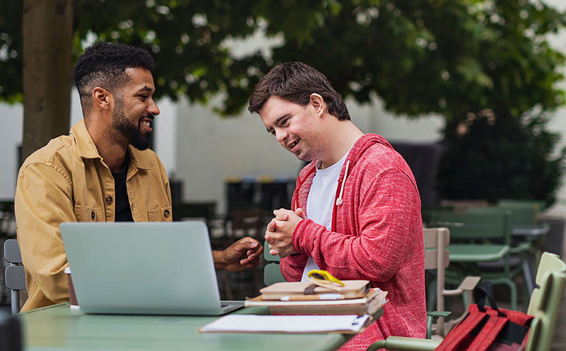 Clinical assessment professional talks with a young man with a disability. They are sitting at an outdoor table with a laptop on top.