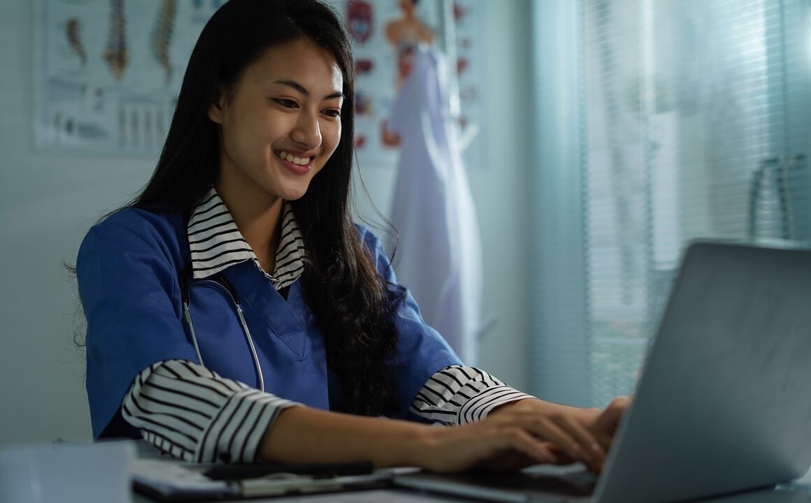 A female doctor in her office uses a laptop to access self-service tools to electronically enroll as a Medicaid provider. She’s wearing a stethoscope around her neck and an anatomy chart and her lab coat are behind her.