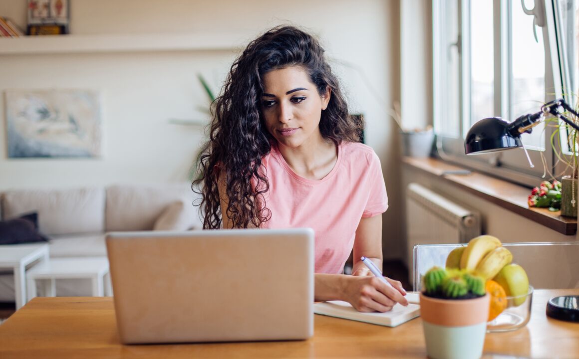 A young woman uses her laptop to chat with a remote agent as she gets help completing an unemployment insurance application. She’s sitting at her desk at home, with a couch behind her and a bowl of fruit next to her. 