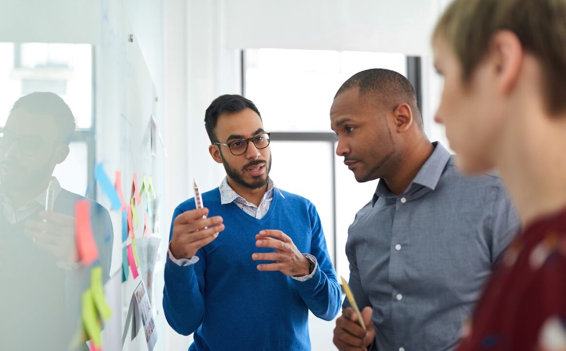 Business process outsourcing expert stands in front of a white board with sticky notes as he explains how to streamline a state health insurance program to his two government clients