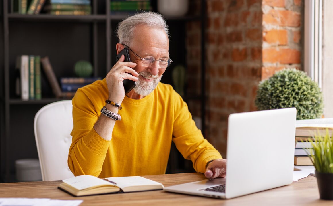 Image of a stylish elderly man smiling and answering phone call while sitting at desk in cozy workplace at home