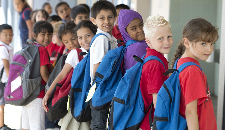 Image of children standing in a line smile over their shoulder with their brand-new backpacks. 