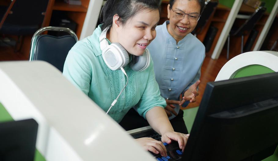 Image of a young blind woman with headphone using a computer with a braille display assistive device while sitting with a senior female colleague. 